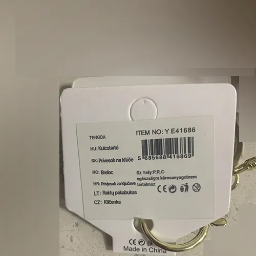 Light brown soft stuffed bear with black plastic eyes and nose, a necklace of small beads on the neck with a metal heart-shaped pendant attached to it. A metal ring and carabiner for hanging are attached to the product.