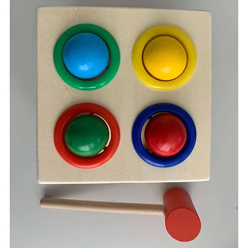 Wooden toy consisting of a box featuring four coloured, circular openings on top, designed to receive coloured (blue, yellow, green, and red) ball-shaped pieces, and a wooden mallet. The toy operates by using the included mallet to drive the ball-shaped pieces into the box. Once inside, the pieces can be retrieved through an opening in the casing and used again.